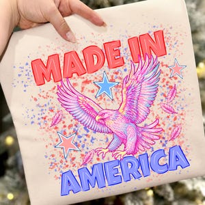 Peut inclure: Un t-shirt beige avec une impression graphique portant le texte MADE IN AMERICA en lettres majuscules rouges et bleues. Le motif comprend un aigle central aux tons roses et bleus, entouré d'éclaboussures de peinture et d'étoiles rouges et bleues.