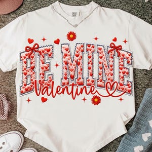 Peut inclure: T-shirt blanc avec l'inscription brodée "BE MINE Valentine" en rouge et blanc. Le motif comprend des cœurs rouges, des nœuds et des motifs floraux, parfait pour la Saint-Valentin.