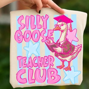 Puede incluir: Un bolso tote beige con la frase "SILLY GOOSE TEACHER CLUB" en rosa. Un ganso de dibujos animados con birrete, gafas y blazer sostiene un libro. Estrellas y rayas azules claras y rosas en el fondo.