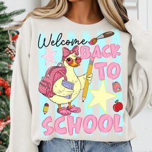 May include: A cream-colored sweatshirt featuring a graphic of a yellow duck wearing pink glasses, a leopard print bow, and a matching backpack. The duck holds a pencil and book. Text reads Welcome BACK TO SCHOOL, accompanied by stars, a paintbrush, and an apple.