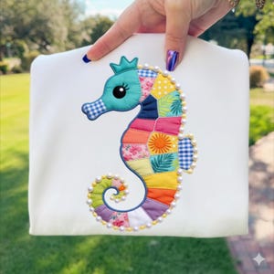 Peut inclure: Sweat-shirt blanc orné d'une applique de hippocampe coloré. L'hippocampe est fait de tissu patchwork aux couleurs vives : bleu, rose, jaune et orange. Il est orné de perles et d'une couronne. Le design est fantaisiste et ludique.