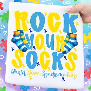 Peut inclure: Tissu blanc avec l'inscription "ROCK YOUR SOCKS" en jaune et bleu, ainsi que deux paires de chaussettes colorées. La phrase "World Down Syndrome Day" est également présente. Le design comprend des rubans bleus et jaunes.