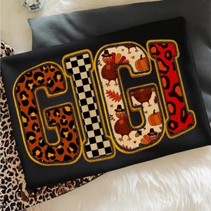 May include: Black sweatshirt with the word "GIGI" in large, colorful letters. The letters feature leopard print, a checkerboard pattern, and Thanksgiving-themed designs with turkeys and pumpkins. The letters are outlined in gold.