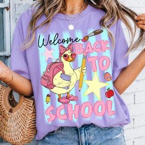 Peut inclure: Un t-shirt violet clair avec un motif représentant un canard jaune portant un nœud rose, des lunettes et des sabots, tenant un sac à dos à motif léopard et un livre. Le design inclut un crayon, un pinceau, une pomme, des étoiles et le texte Welcome BACK TO SCHOOL.