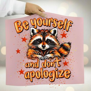 Peut inclure: Une affiche rose avec une illustration de raton laveur et le texte orange "Be yourself and don't apologize". Le raton laveur est marron, noir et blanc, avec des accents orange. Des étoiles rouges et des éclaboussures oranges entourent le raton laveur.