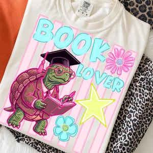 Peut inclure: Un t-shirt crème avec une illustration d'une tortue verte portant une toque de diplômé, des lunettes et un costume marron tout en tenant un livre ouvert. Le texte BOOK LOVER est en bleu clair, avec une étoile jaune et deux fleurs.