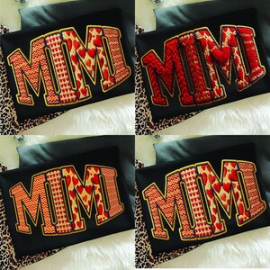 May include: Four black t-shirts with the embroidered word "MIMI" in different styles. The letters are in red, gold, and white, with heart and zigzag patterns. The shirts are on a white and leopard print surface.