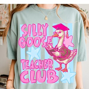 Puede incluir: Camiseta azul claro con las palabras "SILLY GOOSE TEACHER CLUB" en rosa. El dise&ntilde;o presenta un ganso con birrete y gafas, leyendo un libro, con estrellas rosas y azules.