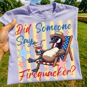 May include: A light purple t-shirt featuring a graphic of a goose wearing a straw hat and sunglasses, sitting in a beach chair. The goose holds a red, white, and blue drink. Text reads: Did Someone Say Firequacker? Background includes stars and stripes.
