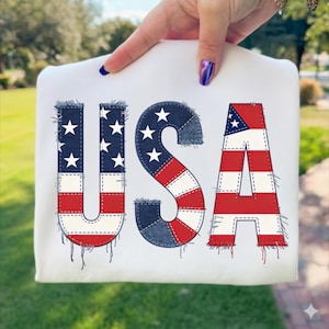 Peut inclure: Sweat-shirt blanc avec le mot "USA" dans un design patriotique. Les lettres sont conçues pour ressembler à du denim et au drapeau américain. Les lettres ont un aspect vieilli avec des bords effilochés.