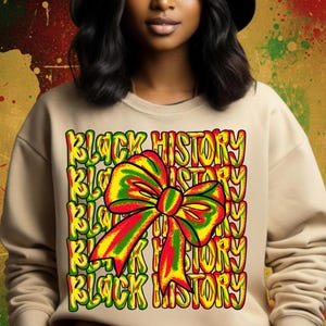 Op de afbeelding: Beige sweatshirt met een kleurrijke afbeelding. De afbeelding bevat de tekst "BLACK HISTORY" in een herhalend patroon, met een grote strik in rood, geel en groen. De sweatshirt heeft lange mouwen en een ronde hals.