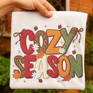 Peut inclure: Un t-shirt blanc avec l'inscription "COZY SEASON" dans un design coloré sur le thème de l'automne. Les lettres sont dans différentes textures et couleurs, dont le rouge, l'orange, le vert et l'imprimé léopard. Le design comprend des feuilles d'automne, des glands et de petites fleurs blanches.