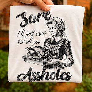 Könnte beinhalten: Weißes T-Shirt mit einer Illustration im Vintage-Stil einer Frau, die einen gebratenen Truthahn hält. Der Text auf dem Shirt lautet "Sure I'll just cook for all you Assholes" in fetter, schwarzer Schrift. Die Frau trägt eine karierte Schürze.