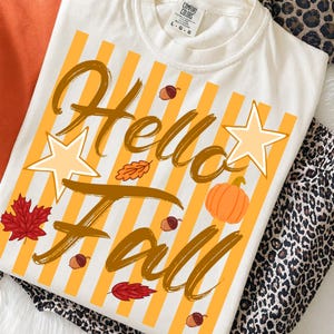 Könnte beinhalten: Ein cremefarbenes Kurzarm-T-Shirt mit einem Grafikdruck aus gelben vertikalen Streifen. Der Text "Hello Fall" ist in brauner Schreibschrift gehalten und von Illustrationen eines Kürbisses, Eicheln, Blättern und zwei hellgelben Sternen umgeben.