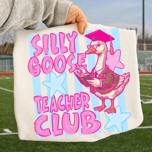 Puede incluir: Un bolso tote crema con un dibujo de un ganso con gafas, birrete y blazer, sosteniendo un libro y un bol&iacute;grafo. El texto "SILLY GOOSE TEACHER CLUB" es en rosa, con estrellas y rayas azules.