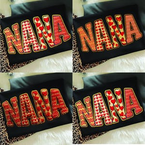 Peut inclure: Quatre t-shirts noirs avec le mot "NANA" brodé dans différents motifs. Les lettres sont rouges, dorées et blanches, avec des motifs de cœurs et de zigzags. Les chemises sont présentées sur une surface blanche et à imprimé léopard.