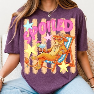May include: Purple t-shirt with a graphic of a lioness wearing sunglasses, a crown, and jewelry, relaxing in a beach chair and holding a cocktail. The word "SPOILED" is in pink letters with star accents.