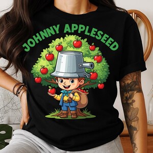 May include: Black t-shirt featuring a cartoon illustration of Johnny Appleseed. The design includes a tree with red apples, a boy with a bucket hat, and the text "JOHNNY APPLESEED" in green.