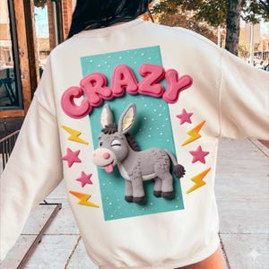 Peut inclure: Sweat-shirt crème avec un design fantaisiste. Le dos présente le mot "CRAZY" en rose, en lettres 3D au-dessus d'un âne gris avec la langue sortie. Des étoiles roses et des éclairs jaunes entourent l'âne sur un fond turquoise.