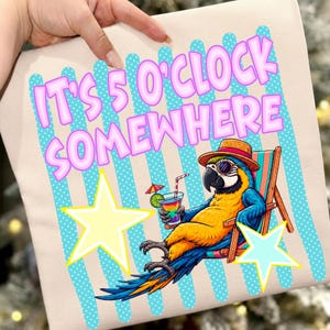 Peut inclure: Un t-shirt beige avec un motif représentant un ara bleu et jaune portant un chapeau de paille et des lunettes de soleil, allongé dans une chaise de plage avec un cocktail coloré. Le texte IT'S 5 O'CLOCK SOMEWHERE est en rose sur des rayures à pois bleu sarcelle.