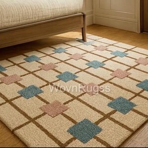 May include: Beige area rug with a brown grid pattern and alternating blue and pink diamond shapes. The rug is placed on a hardwood floor, partially under a wooden bed frame. The rug has the text "WovnRugss".
