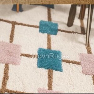 May include: A close-up of a white rug with a geometric pattern. The rug features brown lines forming a grid, with squares of teal and pink. The rug has a soft, textured appearance, suggesting a comfortable and decorative floor covering.