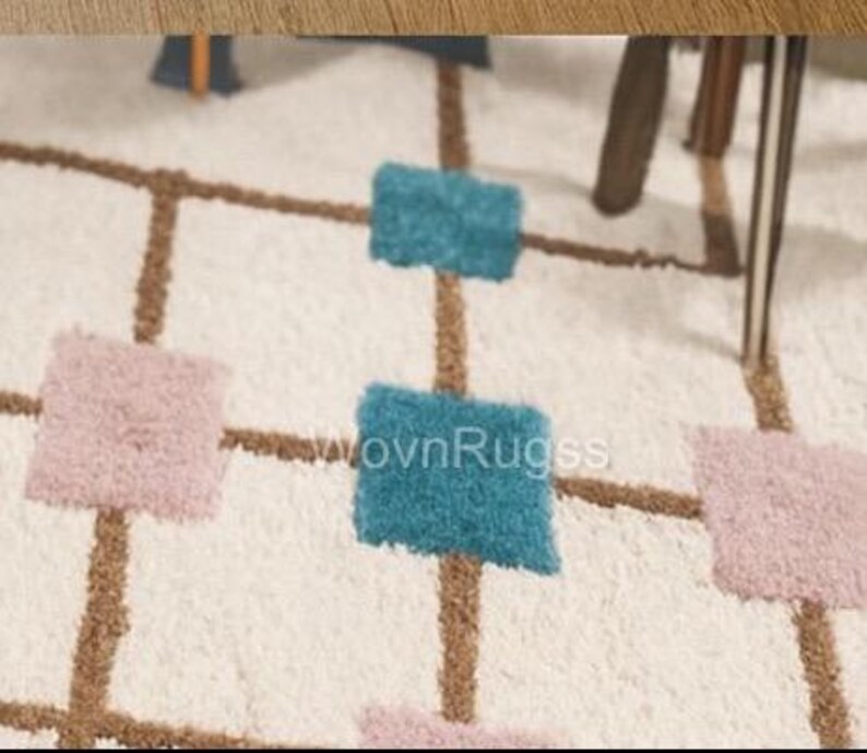 May include: A close-up of a white rug with a geometric pattern. The rug features brown lines forming a grid, with squares of teal and pink. The rug has a soft, textured appearance, suggesting a comfortable and decorative floor covering.