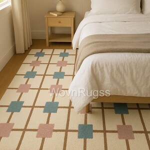 May include: A rectangular area rug with a cream-colored background and a grid pattern of brown lines. The grid is punctuated with alternating squares of blue and pink. The rug is placed next to a bed with white bedding.
