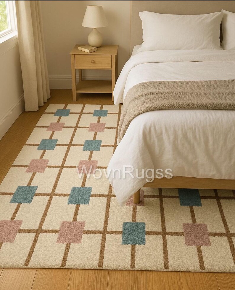 May include: A rectangular area rug with a cream-colored background and a grid pattern of brown lines. The grid is punctuated with alternating squares of blue and pink. The rug is placed next to a bed with white bedding.