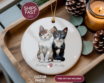 May include: A white ceramic ornament featuring two cat portraits, one with brown and white fur, the other black and white with blue eyes. The names "Harley" and "Misty" are written below the cats. A paw print is between the names.