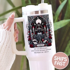 May include: White tumbler with a large handle and a decorative graphic. The graphic features a skeleton reading a book, surrounded by roses and candles. Text on the tumbler reads "SHE SEES RED FLAGS... AND TURNS THE PAGE ANYWAY PROPERTY OF - BELLA."