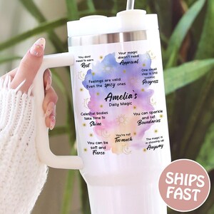 May include: White tumbler with a handle and a lid, featuring a watercolor design with text. The text includes phrases like "Amelia's Daily Magic", "Shine", and "Boundaries". The tumbler has a straw and a silver band.