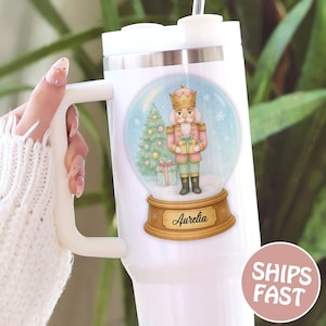 May include: White insulated tumbler with a handle, featuring a snow globe design. The snow globe contains a pink and gold nutcracker, a Christmas tree, and the name "Aurelia". A pink circle with "SHIPS FAST" is visible.
