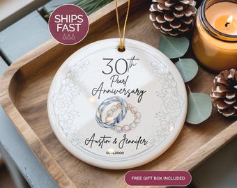 May include: A white ceramic ornament commemorating a 30th Pearl Anniversary. The ornament features a floral wreath design, two intertwined rings, and the names "Austin & Jennifer" with the date 02.11.2000. A gold string is attached for hanging.