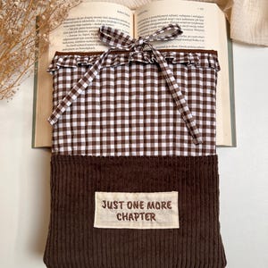 May include: A rectangular book sleeve featuring a brown and white gingham quilted upper section with a ruffled trim and a tie closure. The lower section is made of dark brown corduroy fabric with a rectangular patch reading JUST ONE MORE CHAPTER.