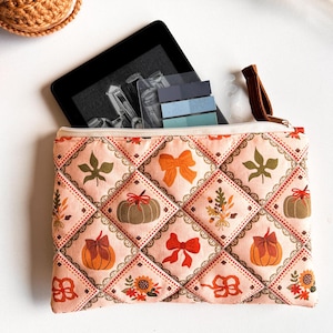 Peut inclure: Une pochette en tissu matelassé avec un fond rose clair présentant un motif en losanges. Chaque losange contient des illustrations de citrouilles orange, de nœuds rouges et orange, de tournesols et de feuilles d'automne. Une tablette noire est insérée.