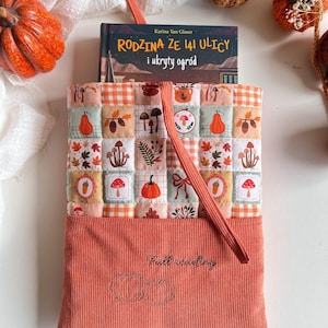 Könnte beinhalten: Eine gesteppte Buchhülle aus Stoff mit einem Patchwork-Muster aus Kürbissen, Pilzen, Eicheln und Blättern in Orange, Creme und Grün. Der untere Teil besteht aus orangefarbenem Kord mit gestickten Kürbissen und dem Text Full reading.