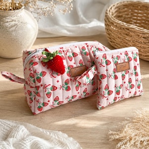 Könnte beinhalten: Eine rosa gesteppte Kosmetiktasche und ein passendes kleines Täschchen, beide mit einem sich wiederholenden Erdbeermuster verziert. Eine gehäkelte Erdbeere ist an der Tasche befestigt. Die Taschen haben einen weißen Reißverschluss und ein kleines braunes Etikett.