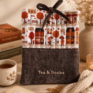 May include: A quilted fabric book sleeve featuring a white background with a repeating autumn town scene including trees, buildings, and people with umbrellas. The bottom section is dark brown corduroy with the embroidered text Tea & Books.