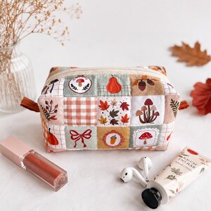 Op de afbeelding: Een rechthoekig stoffen etui met een patchworkontwerp met herfstthema-illustraties, waaronder pompoenen, paddenstoelen, eikels, bladeren en een rode strik. De tas heeft een beige ritssluiting en een zachte, doorgestikte textuur met een ruitpatroon.