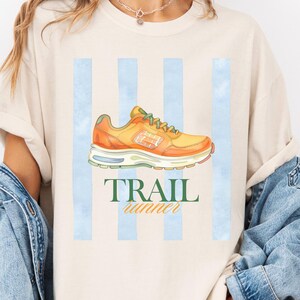 May include: A beige short-sleeve t-shirt featuring a graphic of an orange running shoe centered over three vertical light blue stripes. Below the shoe, the text TRAIL runner is printed in dark green and orange lettering on the front of the shirt.