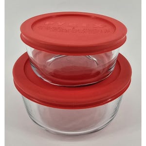 Puede incluir: Dos cuencos de vidrio transparente Pyrex con tapas rojas brillantes. Los cuencos están apilados, con el cuenco más pequeño encima del más grande. Las tapas tienen el logotipo y el texto de Pyrex. Ideal para guardar alimentos.