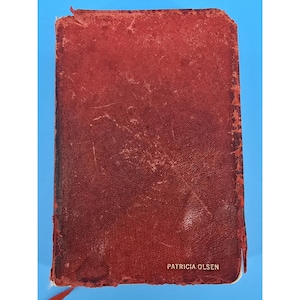May include: A small, worn, red book with the name "PATRICIA OLSEN" printed in gold at the bottom. The cover shows signs of wear and tear, with visible creases and scuffs. The book has a slightly distressed appearance.