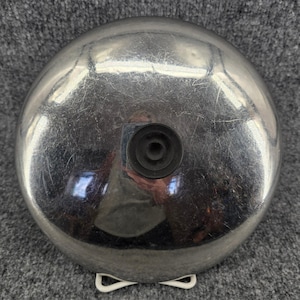 May include: A circular, polished silver-tone metal dome with a smooth, reflective surface. A small, black, circular rubber knob is centered on the top of the dome. The metal surface shows fine, light scratches across its entire exterior.
