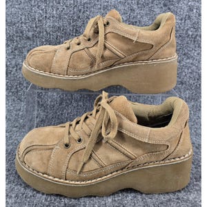 May include: A pair of tan suede platform sneakers featuring a thick, chunky rubber sole, matching tan laces, and reinforced stitched paneling across the upper. The shoes have a rounded toe design and a padded collar for ankle support.