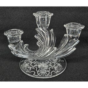 Può includere: Un candelabro in vetro trasparente con tre portacandele. Il candelabro presenta un design decorativo a spirale e poggia su una base rotonda con dettagli floreali e a nastro incisi. I portacandele sono circolari.