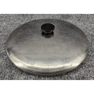 May include: A circular, brushed metal pot lid with a smooth, slightly domed surface. The lid features a small, centered, black circular knob handle made of a solid material, providing a simple grip for lifting the lid from a cooking vessel.