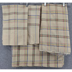 Op de afbeelding: Verschillende beige geruite stoffen, mogelijk beddengoed of gordijnen, worden getoond. Het ruitpatroon heeft dunne lijnen in rood, blauw en geel, waardoor een klassiek, neutraal ontwerp ontstaat. De stof hangt aan metalen hangers.