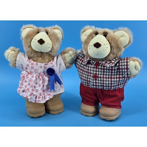 May include: Two plush teddy bears, one wearing a floral dress with a blue ribbon, the other in a plaid shirt and red pants. Both have tan shoes and are set against a blue background.