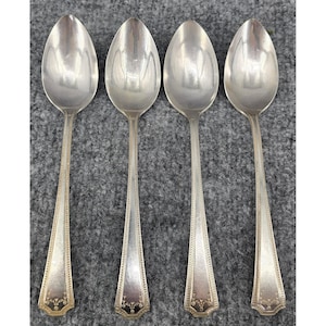 May include: Four silver-toned metal spoons arranged in a vertical row. Each spoon features a smooth, oval-shaped bowl and a slender handle with a decorative beaded border along the edges and a small floral engraving at the base of the handle.